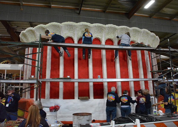 Volunteers on scaffolding glue white petal trim atop a ""Forum" float