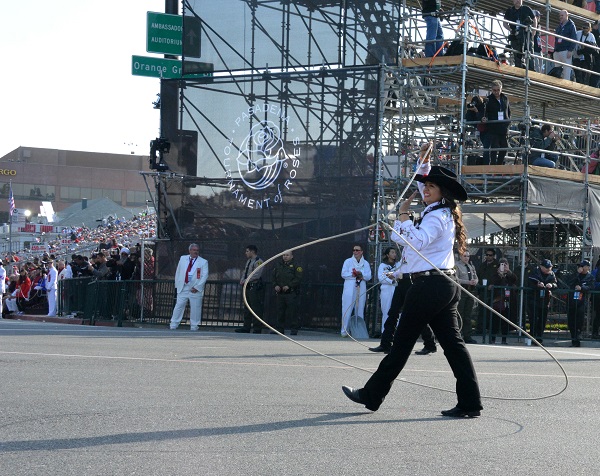 Woman in Western attire and black cowboy hat walks along Colorado Boulevard inside a rope lariat as she twirls it