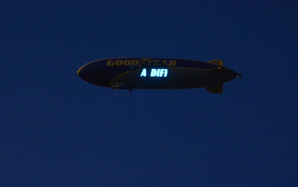 Goodyear blimp in predawn sky flashing letters, "A DIFF"