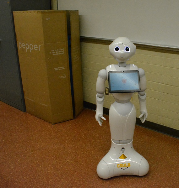 White "Pepper" humanoid robot stands on carpet near a box with her name on it