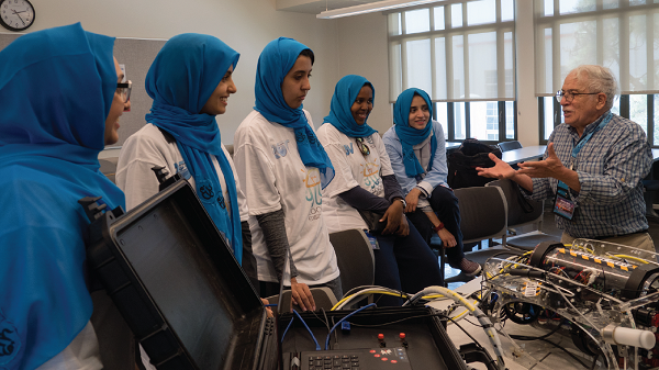 Students in blue hijabs listen as a mentor explains how to create a robot for MATE Center's underwater robotics competition