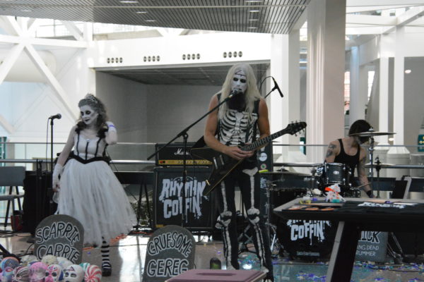 Three "Rhythm Coffin" musicians in dead-white makeup sing in lobby of LA Convention Center, next to "tombstone" signs with their names