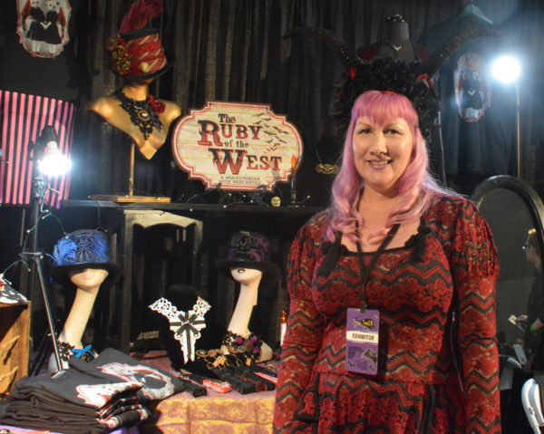 Smiling vendor in horned headgear stands near her booth of "Ruby of hte West" merchandis at ScareLA 2018