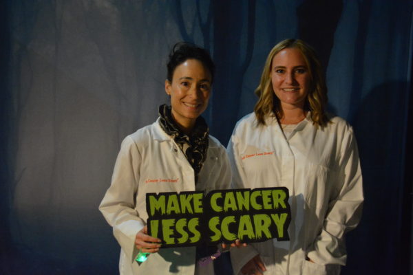 two women in white uniforms with "Make Cancer Less Scary" sign in green lettering at ScareLA 2018