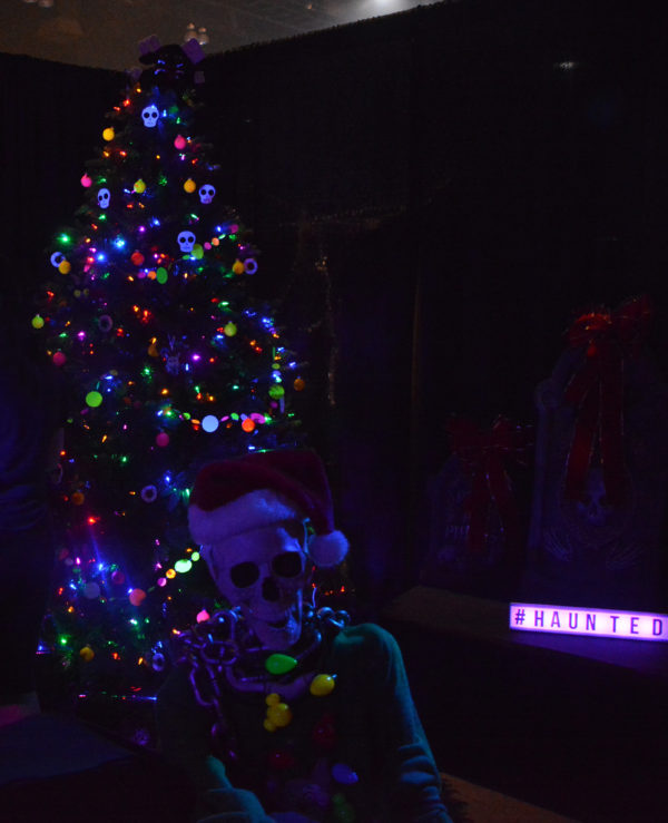 Skeleton in a Santa hat sits next to a Garvin Unliited Christmas tree witha "Ghoulish Garland" of skulls and Halloween symbols