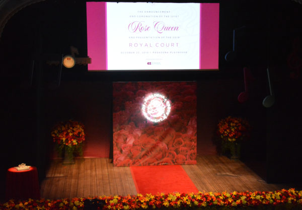 Pasadena Playhouse stage set up for coronation of 101st Rose Queen with red carpet, white Tournament of Roses circular logo and crown on table