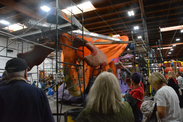 Visitors photograph a Rose Parade® dragon float behind scaffolding with their phones as volunteers work in the background