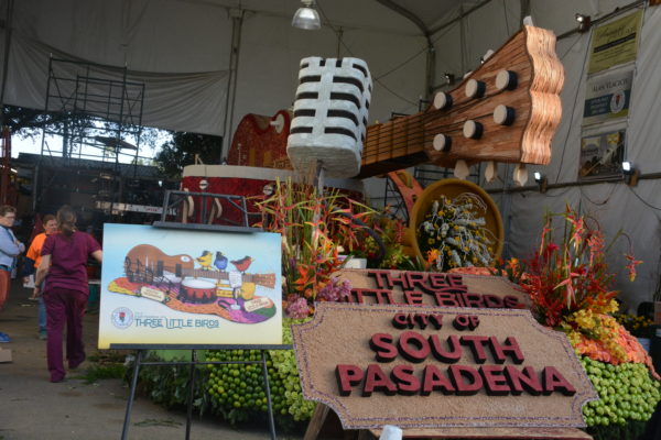 "Three Little Birds" Rose Parade® float in its barn, with rendering and sign, "City of Sauth Pasadena" and an outsized guitar running the length of the float