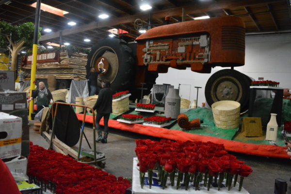 Chipotle Rose Parade® float, with its tractor at the front, stands near boxes of fresh roses and vegetables for decoration