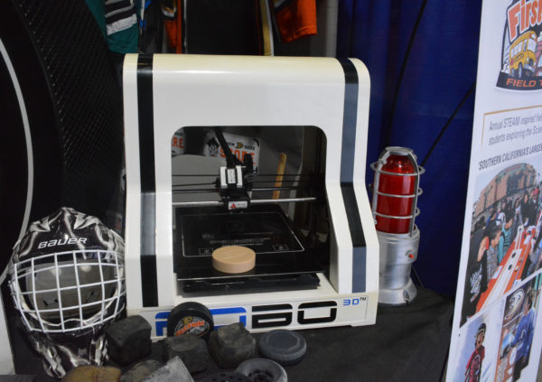 3D printer prints a puck at S.C.O.R.E. exhibit, surrounded by similarly printed pucks and a helmet with designs