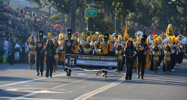 Band in black and gold uniforms with dancers in gold glittery costumes and tubas in the background marches up behind a black banner that reads, "Alabama State University Mighty Marching Hornets"