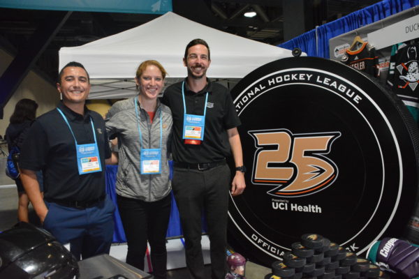 S.C.O.R.E. Program exhibit at 2018 California STEAM Symposium, as Fan Development Marketing Coordinator Gabriel Alamillo, Fan Development Marketing Manager Molly Schaus and Fan Development Marketing Manager Jason Cooper smile from in front of a gian black "puck" with "25" on it in gold.