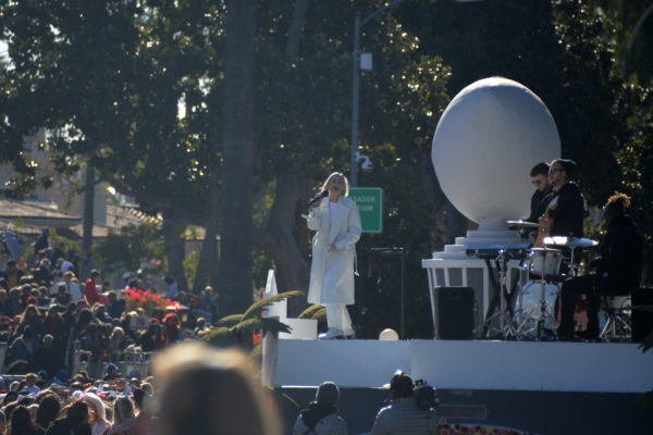 Anne Marie wears white slacks and long jacket as she sings onstage before Rose Parade crowd with two musicians in the background