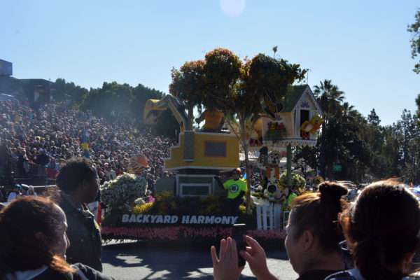 "Backyard Harmony" float with surprised bird in birdhouse, cat in hard hat and bulldog in hard hat operating bulldozer as spectators photograph it with their phones