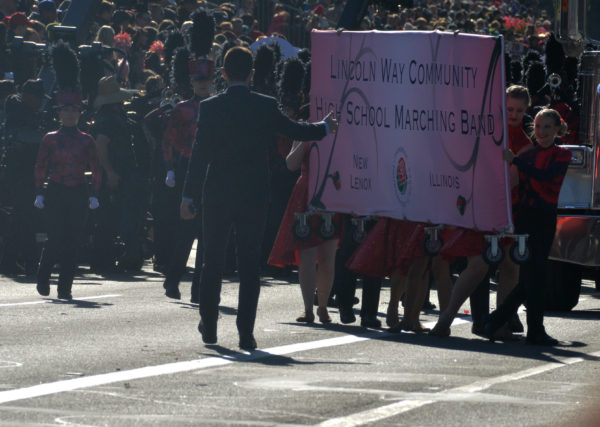 Bandmembers in red shirts and black trousers carry pink sign with "Lincoln Way Community High School Marching Band"