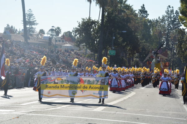 Band members, in white uniforms with gold plumes, march behind a banner reading "Banda Municipal de Acosta"