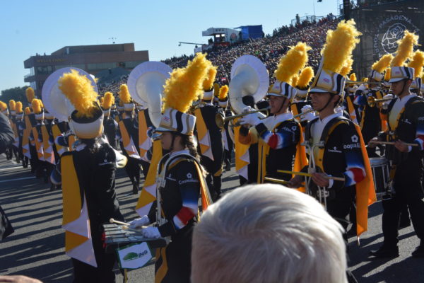Glockenspiel player in navy band uniform with white hat and gold plume plays facing audience directly as column before her heads away at a 45-degree angle