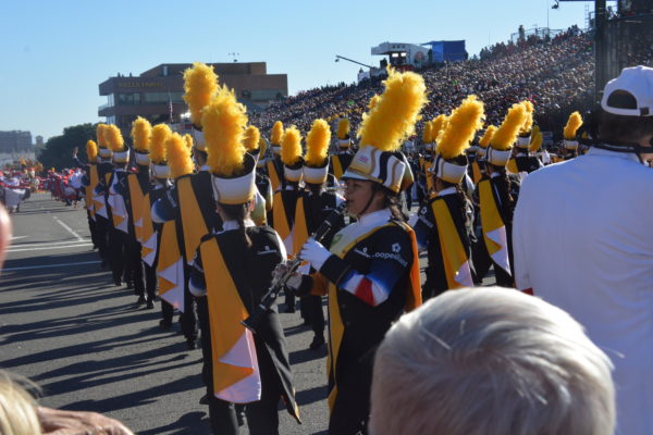 Piccolo player in navy band uniform with white hat and gold plume plays facing audience directly as column before her heads away at a 45-degree angle