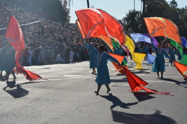 Women in calf-length sea-green dresses with white leis around their necks wave two flags each, of varying colors: orange, blue, yellow, green, purpleand red