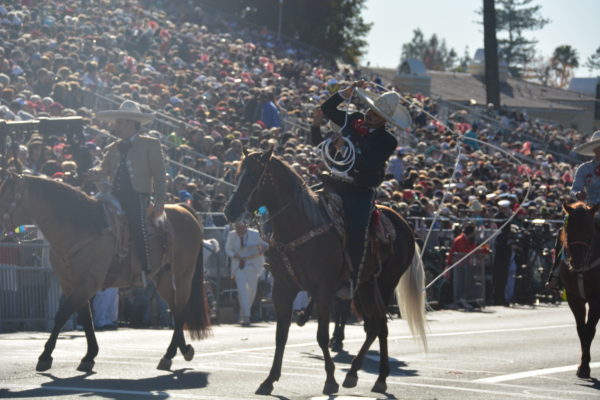 Closeup of Charro rider on bay horse, flanked by two other riders, as he spins a rope lasso while Rose Parade audience looks on from the stands