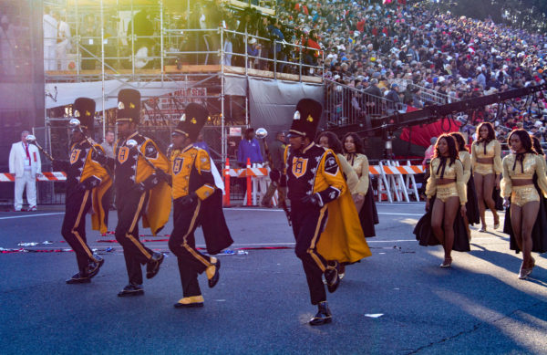 Three African=American drum majors in black uniforms with gold capes march, keeping time with their batons, followed by female African-American marchers in sparkly gold midriff outfits