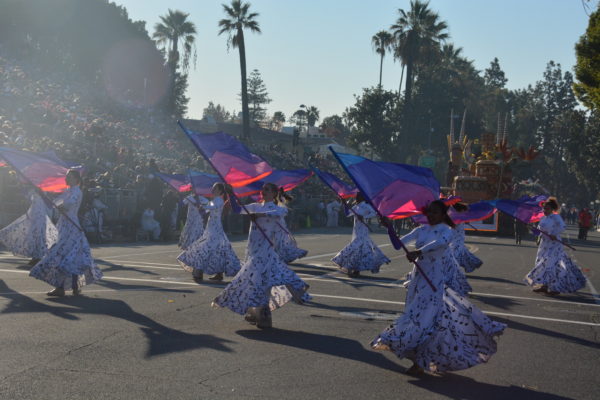 Women in swirling white skirts with purple trim wave pink and purple flags
