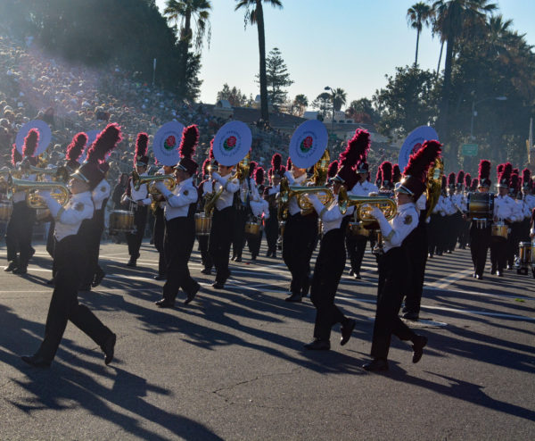 Bandmembers in white shirts and black pants and vests with red-plumed hats play trumpets as they march