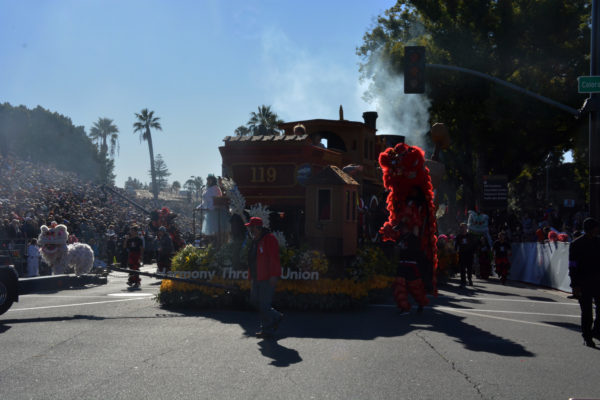 Front view of "Harmony Through Union" float with white lion dancer on one side and red one on another and costumed woman in a white outifit wavinf from the front as the float is towed with palm trees and blue sky in the background