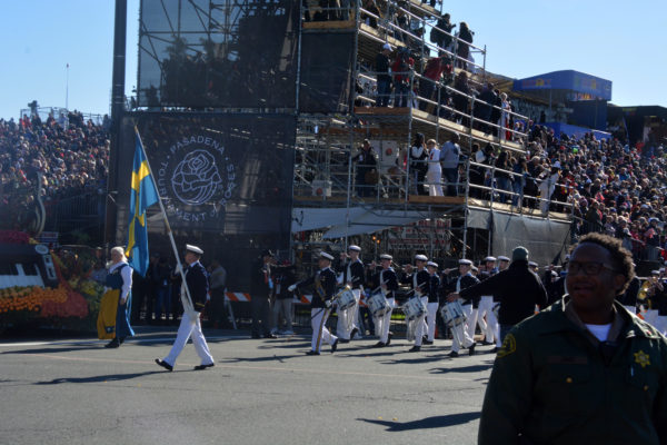 Swedish band, in white trousers and caps with navy-blue jackets, marches ahead of Tournament of Roses symbol on gray wall, following carrier with a Swedish flag