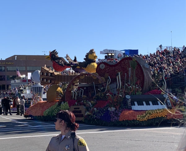 Three Little Birds float shows red guitar in center with three brids perched atop it and keyboard at the back of the float, as it stands on Colorado Boulevard and a female sheriff looks on