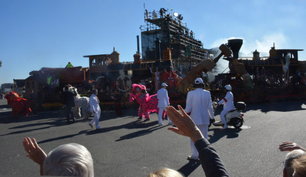 Paradegoers wave as dual-train "Harmony Through Union" float, with hands driving a hammer onto a golden spike, gets towed by with Asian lion dancers dancing alongside it