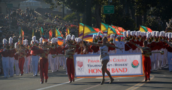 Drum majorette in a sparkly white leotard spreads her arms and smiles in front of a banner that says, "Pasadena City College Tournament of Roses Honor Band", flanked by bandmembers in red slacks and white jackets and with multicolored flags in the background