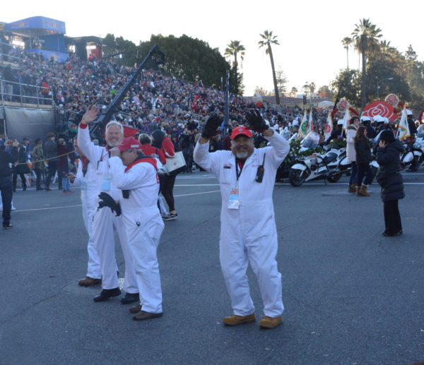 Three float mechanics in white uniforms and red caps smile and raise their hands in the air against background of filled stands and motorcycle officers ready to go