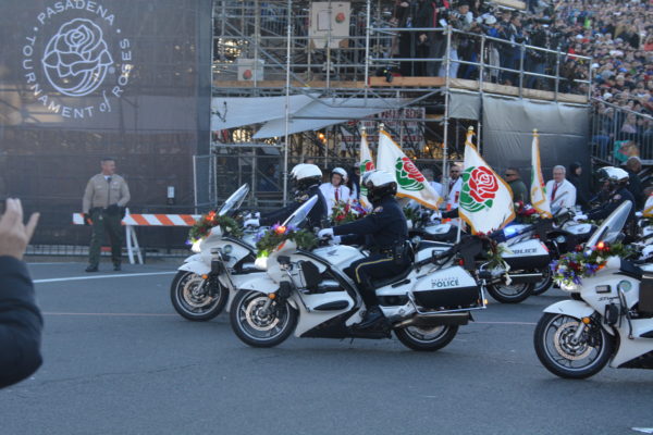 Motorcycle police ride past Tournament of Roses logo on reviewing stand with fluttering white flags with a red rose logo with green leaves on the back of their cycles