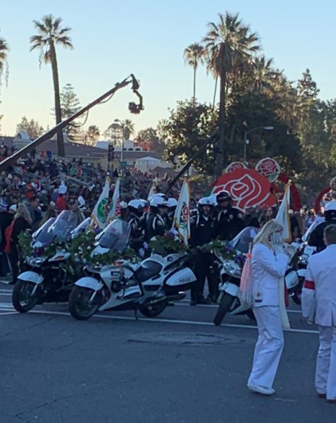 Helmeted motorcycle police stand ready to mount their vehicles at start of parade route as White Suiters confer and rose logo stands in background