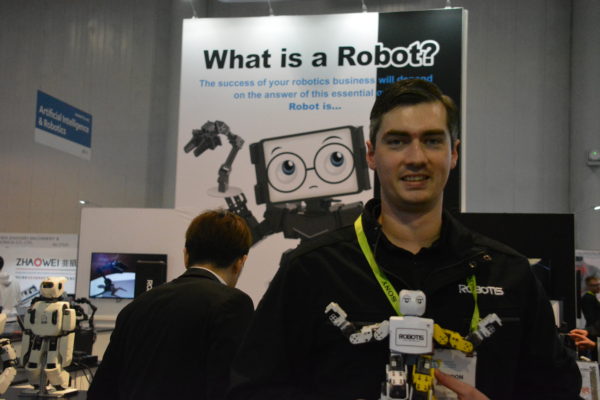 Brandon Antille of Robotis holds up miniature white humanoid robot near "What Is a Robot?" sign