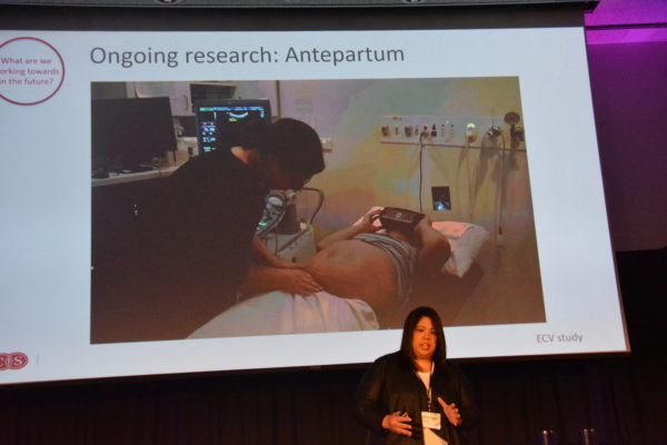 Dr. Wong before a slide of a doctor examining a pregnant woman wearing a VR headset during Virtual Medicine conference