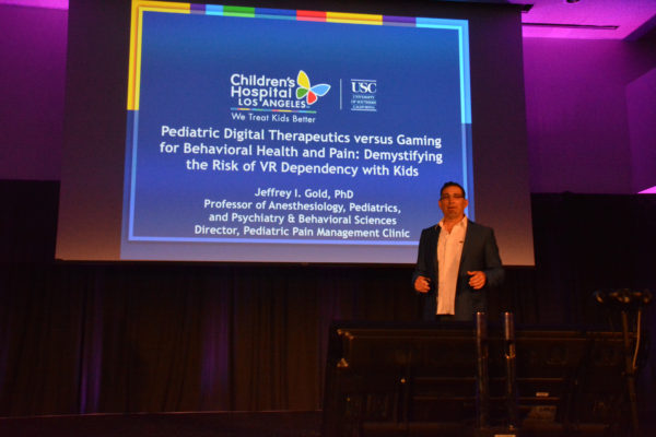 Dr. Jeffrey Gold stands onstage before blue slide with "Pediatric Digital Therapeutics Versus Gaming for Behavioral Health and Pain: Demystifying the Risk of VR Dependency with Kids" title on a blue background during Virtual Medicine 2019
