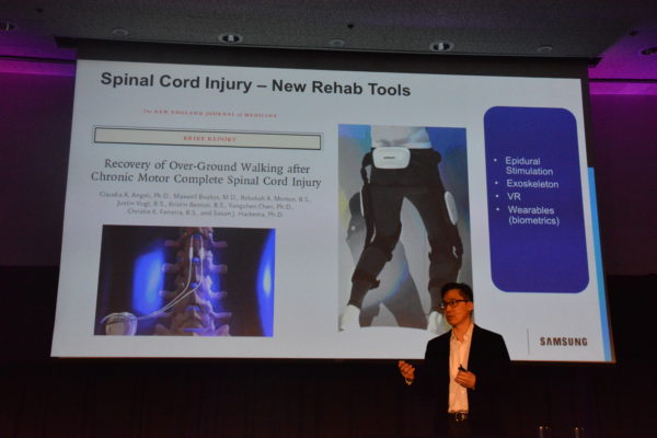 Dr. Rhew in front of a slide showing an "exoskeleton" that allows paraplegic to regain use of legs at Virtual Medicine 2019