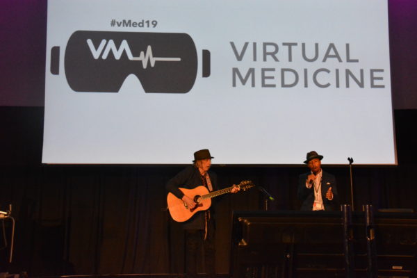 Harmon Clarke sings with guitarist accompanying him near a slide that says "Virtual Medicine"