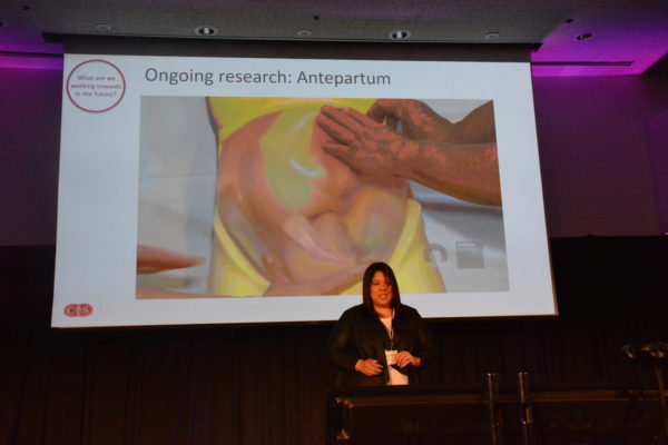 Dr. Melissa Wong stands before slide of doctor palpating a woman's abdomen during Virtual Medicine 2019 conference