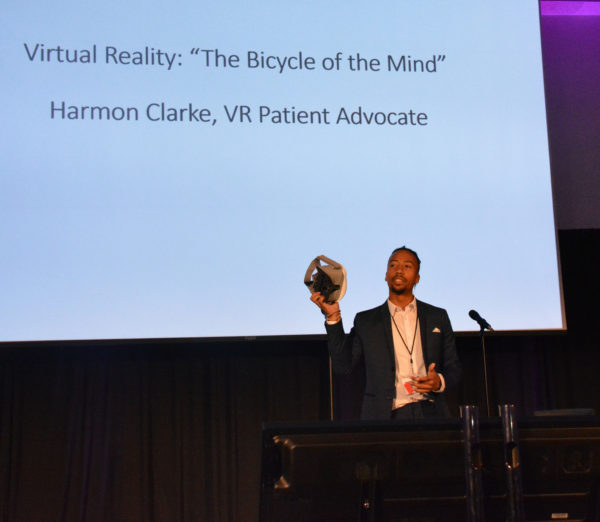 Harmon holds up a VR headset as he stands near a slide that reads: "Virtual Reality: The Bicycle of the Mind, Harmon Clarke, VR Patient Advocate"