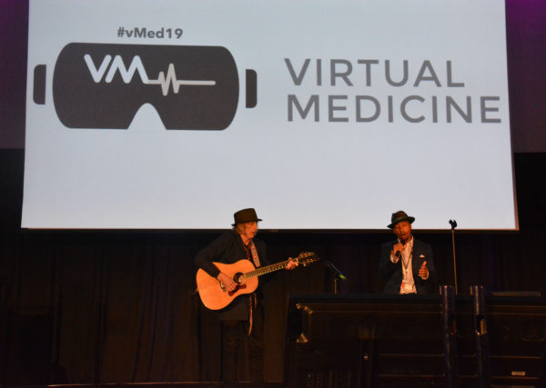 Harmon, wearing a fedora hat, sings to the accompaniment of an acoustic guitar in front of a screen that says, "Virtual Medicine"