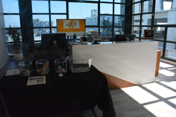 Lobby of Soylent Innovation Labs with food products displayed on a table for visitors and a huge "MakerWalk LA 2019" sign on a video screen before a window showcasing the L.A. skyline