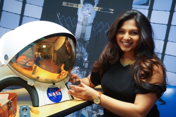 NASA SUITS researcher Paromita Mitra smiles as she poses next to a spacesuit helmet with the blue "NASA" logo