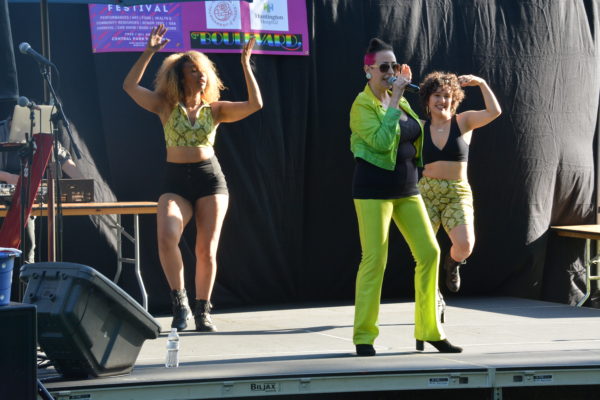 Betsy Body on mic onstage flanked by two jazz dancers