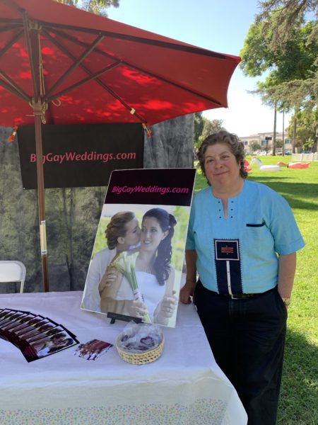 Susan Goldman smiles as she stands next to her table with its Big Gay Weddings logo and photo of a couple kissing at their wedding