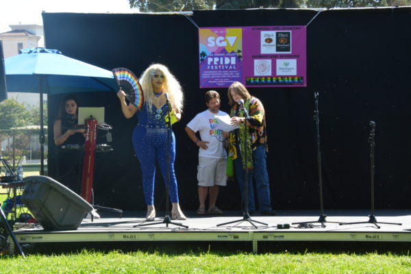 MC Clitdreese holds rainbow fan as Reverend Pat and Reverend Lee speak in background at San Gabriel Valley Pride