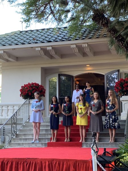Six finalists stand on either side of steps as female White Suiter escorts another arrival