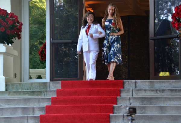 Female White Suiter with red tie escorts first finalist on her arm down the red-carpeted stairs